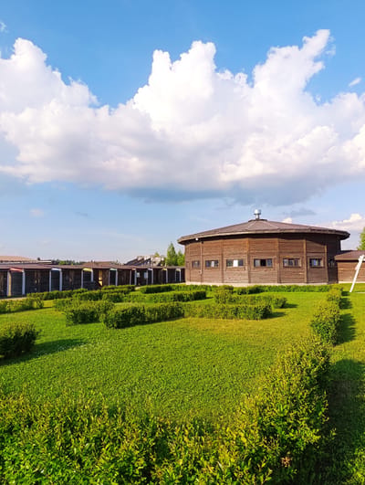 The image shows an outdoor scene in the city of Ivanovskoe, Russia. In the background, there is a large, circular, wooden building with a brown, sloped roof and several small, rectangular windows. In front of this building, there is a row of smaller, wooden structures with corrugated metal roofs, each with a different colored panel on the front. The foreground is dominated by a lush green lawn with neatly trimmed hedges arranged in rectangular patterns. The sky above is a clear blue with fluffy white clouds. The scene appears to be a recreational or park area.