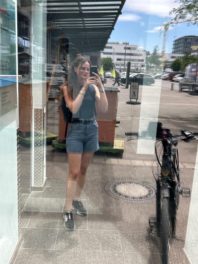 The image is a full-body mirror selfie taken by a young woman standing on a tiled sidewalk in the city of Heilbronn, Germany. She is wearing glasses, a dark sleeveless top, denim shorts, and black and white sneakers. She has long, wavy brown hair and is holding a smartphone in her right hand, taking the photo. A black backpack is visible on her shoulders.

The reflection in the glass shows a street scene with parked cars, a bicycle in the foreground, buildings, a crane, and a large banner with text including "KÖLLE ZOO" and "MEHR". A man is walking in the background on the left side of the image. The ground is paved with gray tiles and a circular metal manhole cover is visible. The overall environment appears to be an urban commercial area.