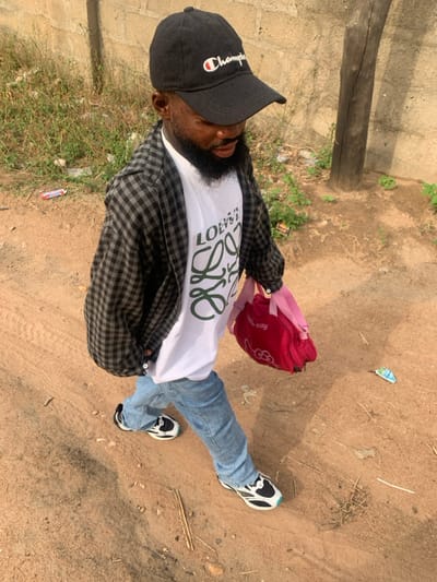 A dark-skinned individual, appearing male, with a full dark beard, is depicted walking on an unpaved dirt path in Eika-Adagu New Layout, Nigeria. He wears a black baseball cap with a white and red "Champion" logo, a white t-shirt featuring a green stylized "LOEWE" graphic, an unbuttoned black and grey plaid long-sleeve shirt over the t-shirt, light blue denim jeans, and black, white, and light blue athletic shoes. In his left hand, he carries a red bag with pink straps and a white "Hello Kitty" logo. His right hand is placed inside his pants pocket. The individual's head is tilted downward, and his gaze is directed towards the ground. The immediate environment consists of dry, light brown earth, scattered small pieces of debris, and sparse green vegetation. A light-colored concrete block wall is visible in the background, with a weathered dark wooden post or tree trunk positioned to the right of the individual. The scene is illuminated by natural daylight, casting shadows to the right of the subject.