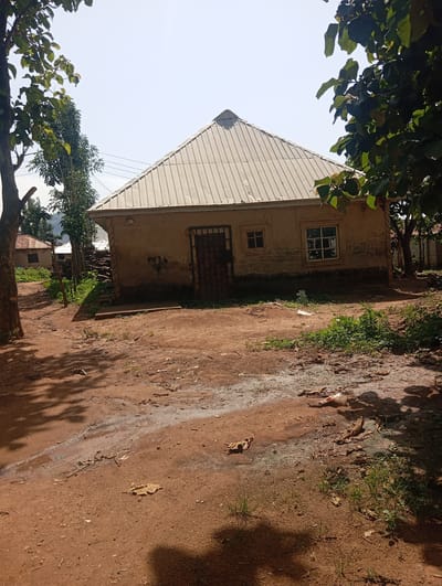 In Karsana 2, Nigeria, a light brown, one-story building with a corrugated metal roof stands on a dirt path. The building has a dark metal door and two windows. One window, to the left of the door, is small and square with a light frame. The other, to the right of the door, is larger and rectangular with a white frame. The walls of the building are adorned with dark graffiti. The ground in front of the building is uneven, with patches of standing water and scattered debris, including dried leaves and a plastic bag. Trees with green leaves surround the building and path, casting shadows on the ground. In the background, another building with a reddish-brown roof is visible, along with vegetation and what appears to be a distant hillside. The sky is clear and bright.