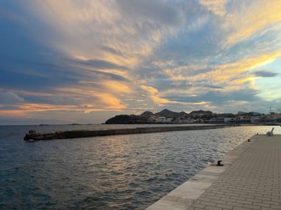 This image depicts a coastal scene in Cabo De Palos, Spain, during either sunrise or sunset. The sky, covering the upper portion, presents a gradient of deep blue on the left, transitioning to orange and yellow hues towards the right, dominated by wispy, elongated stratiform clouds. The water, occupying the lower half, displays gentle ripples and reflects the sky's colors.
In the mid-ground, a concrete breakwater or pier extends horizontally from the left, its left end partially submerged among visible rock formations. To the right of the breakwater, a coastal town is visible, featuring numerous light-colored buildings clustered at the base of undulating, dark-toned hills in the distant background.
The immediate foreground on the right consists of a paved quay or dock, surfaced with light-grey rectangular pavers. Several dark-colored mooring bollards are anchored along its edge, and a white bench-like structure is partially visible at the far right. No people or specific activities are present.