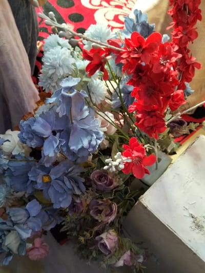 A floral arrangement composed of artificial blue and red flowers, along with some muted pink and purple roses, is displayed. The arrangement is partially obscured by a person's leg covered in a light pink garment and a dark denim fabric. A red and black patterned rug is visible in the background. The scene appears to be indoors, possibly in a residential setting in Cottrueng, Indonesia. The lighting is moderate, and the focus is on the flowers.