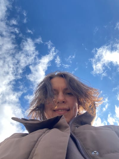 A low-angle, eye-level shot shows a person with short, light brown hair and a brown jacket, taken against a partly cloudy blue sky. The individual is smiling slightly. The setting does not appear to be Nizhnie Yaki, Russia.
