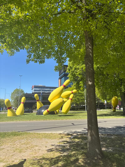 The image depicts a roadside scene in Eindhoven, Netherlands, featuring a public art installation of oversized yellow bowling pins with red stripes at their necks. The pins are arranged as if struck by a bowling ball, which is also part of the sculpture. The installation is set on a grassy area adjacent to a paved road, with trees providing shade. Buildings can be seen in the background, under a clear blue sky.