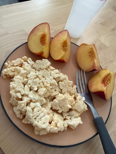 A close-up, overhead shot shows a plate of scrambled eggs and sliced peaches on a wooden table. A white plastic cup is in the upper right corner. The plate is tan with a dark blue rim and contains a generous portion of fluffy, pale yellow scrambled eggs. Four slices of peaches are arranged around the eggs. The peaches are a vibrant yellow with reddish-pink skin and show their seed cavities. A silver fork with a dark gray handle rests on the right side of the plate, partially submerged in the scrambled eggs. The wooden table has a light, natural grain. The scene appears to be an indoor setting, possibly a home or cafe, and there is no specific information to indicate its location in Almada, Portugal.