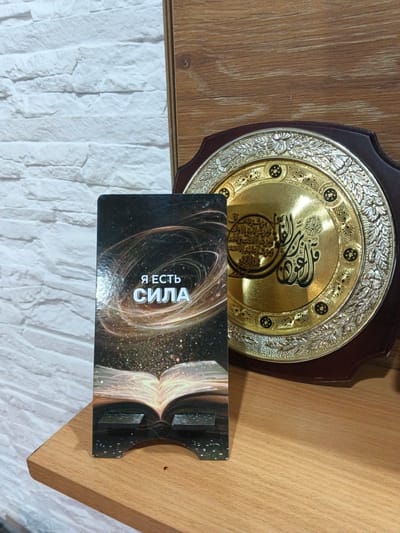 A close-up, eye-level, interior shot focuses on a wooden shelf, likely in Novyy, Russia, displaying two distinct objects. On the left, a vertical, dark-colored stand holds an open book with a cosmic, starry background. Text in white Cyrillic script, "Я ЕСТЬ СИЛА" (I AM POWER), is prominently displayed on the book's cover. To the right of the stand, a large, ornate, circular golden plaque with intricate silver detailing is mounted on a dark wooden base. The plaque features Arabic calligraphy and decorative floral motifs. The background consists of a white, textured brick wall on the left and a wooden surface on the right, suggesting a home or office environment. The lighting is moderate, casting soft shadows and highlighting the textures of the objects and surfaces.