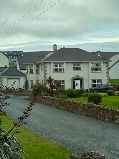 The image shows a large, two-story house in Dunfanaghy, Ireland, with a gravel driveway leading up to it. The house is painted white with stone accents and has a dark gray roof. There are several windows on the front of the house, some of which are open. A dark-colored SUV is parked in the driveway. To the left of the main house is a smaller, attached garage with a roll-up door. The property is surrounded by a low stone wall and a well-maintained lawn with various shrubs and plants. In the foreground, there are some dark-colored plants with red flowers. The sky is overcast.