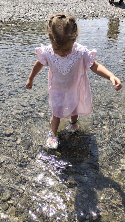 A young girl, seen from a high angle, stands in shallow, clear water. She is wearing a pale pink dress with white lace detailing around the neckline and cap sleeves, and what appear to be white sandals with colorful floral accents. Her hair is pulled up in several small ponytails. The water is shallow enough to reveal a rocky, pebbly riverbed, and sunlight creates shimmering reflections on the water's surface. The girl's shadow is cast on the riverbed to her right. The background consists of more pebbles and a blurred shape in the upper right corner, possibly a person or object. This scene likely depicts a child playing in a river or stream in a natural or semi-natural outdoor environment.