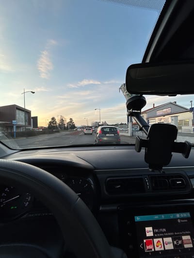 The image depicts the interior of a car looking out through the windshield. A rearview mirror and a phone mount are attached to the glass. Outside, a street is visible with a line of cars waiting in traffic and buildings. A radio display shows "FM/FUN" and "96.30 MHz Fun Radio." The speedometer is partly visible with markings.