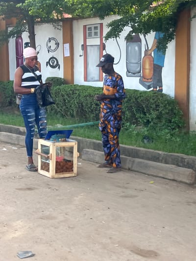 This outdoor street scene, set in Ijegun, Nigeria, features two individuals on a light-colored concrete or dirt path, under bright daylight conditions.

In the foreground, a person stands beside a small, wooden and glass vending cart. This individual wears a black and white striped sleeveless top, distressed blue jeans, a light pink headwrap, and dark sandals. They are holding a black plastic bag in one hand. The vending cart contains numerous brown, spherical items, likely fried snacks or street food.

Further back, another person stands, oriented towards the left. They are dressed in a full-body outfit with a distinctive pattern of blue, purple, and orange or yellow leaf-like shapes, and wear a dark cap or hat.

Behind the individuals, a white building wall is visible, partly obscured by green bushes and trees. The wall features murals that depict gas-related themes: one shows a person holding a large orange gas cylinder with a grey cylinder nearby, and another displays illustrations of gas regulators and burner components. A painted window is also visible on the wall. The presence of these murals suggests the location may be near a gas cylinder sales or refill point, complementing the informal street vending activity. No clear text is discernible on the wall or vending cart.