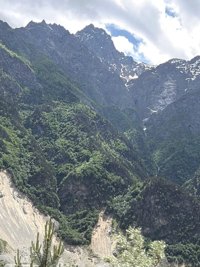 The image depicts a mountainous landscape near Buron, Russia, characterized by steep, rocky peaks interspersed with dense green forests. The terrain is rugged, with visible rock faces and scree slopes, some of which appear to have been affected by landslides. Patches of snow can be seen on the higher elevations, suggesting a high-altitude environment. The sky is partly cloudy, with sunlight breaking through in places, illuminating the slopes. The foreground shows some foliage, possibly trees and bushes, indicating a natural, untamed environment. The scene conveys a sense of wilderness and the raw power of nature.