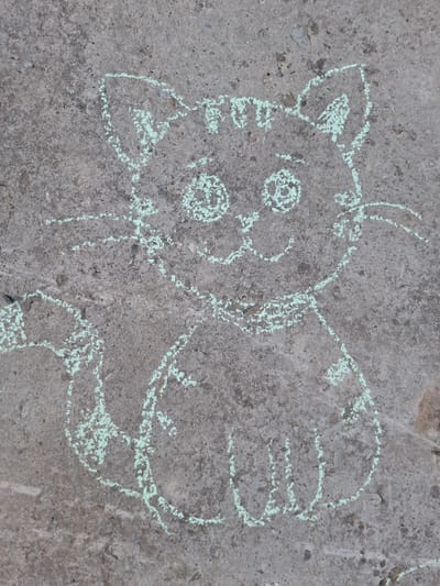 A chalk drawing of a cat-like figure is on a concrete surface. The location is uncertain, but the city is Амзя, Russia. The figure is a light green color and has a roughly circular head with two pointed ears, two round eyes, a small nose, and a mouth. There is a tail on the right side of the figure. There are 2 antennas above the cat's head.