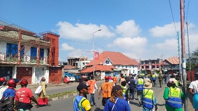 The image displays a daytime urban street scene in Gisenyi, Rwanda, under a clear blue sky with scattered white cumulus clouds. A group of approximately 30-40 individuals, largely adults, is observed walking away from the camera in the right lane of an asphalt road. Many individuals wear high-visibility vests: some are orange with white text or numbers (e.g., "527"), others are green (some with "CUSTOM" visible), and some are yellow and blue. Several individuals in the foreground and mid-ground wear red helmets, and one individual wears a red t-shirt with "JOIN US" text.

Buildings line both sides of the street. On the left, a two-story white building with blue-tinted windows features red external metal balcony structures or fire escapes. Its ground floor exhibits several closed storefronts. Beyond it, a blue pickup truck with red objects in its bed is parked. In the mid-ground, a prominent building with a red-tiled roof and white facade, including a distinct gabled section, stands on the right. A street light pole with a red flag or sign bearing a white "V" logo is positioned in front of this building. Further down the street, additional multi-story commercial and residential structures are visible, some appearing partially constructed or aged. Overhead utility wires crisscross the frame, connecting various poles, including a tall, weathered utility pole with a blue band on the far right. The road features a painted median strip, and sparse vegetation is visible on the far right.