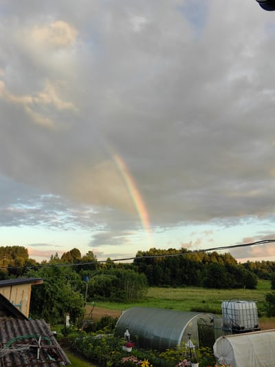 The image depicts an outdoor rural scene with a prominent rainbow arcing across a cloudy sky. In the foreground, there are several structures, including a greenhouse with a white water tank next to it, a partially constructed building with visible insulation, and a section of a corrugated roof. The middle ground features a lush green landscape with a dense line of trees bordering a field. The background is dominated by the sky, which is filled with a mix of grey and lighter, possibly sunlit, clouds. The lighting suggests it is either early morning or late afternoon. The scene appears to be located in a rural area, possibly a village or a dacha settlement. The location is identified as Дзямідавіцкі сельскі Савет, Belarus. No people are visible in the image.