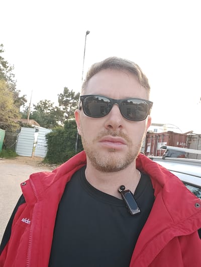 An adult male is depicted in a close-up, outdoor selfie-style photograph. He has short, light brown hair, a trimmed light brown beard, and wears black square-framed sunglasses that reflect a vehicle and a phone. He is dressed in a black crew-neck shirt, over which he wears an unzipped red hooded jacket with "sida" partially visible on the left sleeve. A black electronic device, possibly a Bluetooth earpiece or portable speaker, with a dark rectangular screen, is clipped to the collar of his black shirt.
The scene is set in Nesher, Israel. To the subject's left, a dirt path leads towards a green metal fence post and light-colored corrugated metal fencing, partially open. Dense green foliage and trees are visible beyond the fencing. To the subject's right, the roof and roof rack of a silver vehicle are visible, behind which multiple rusty red shipping containers are present, some bearing white markings including "3EBBE". A tall, slender light pole extends upwards on the right side of the background. The sky is bright and slightly overexposed.