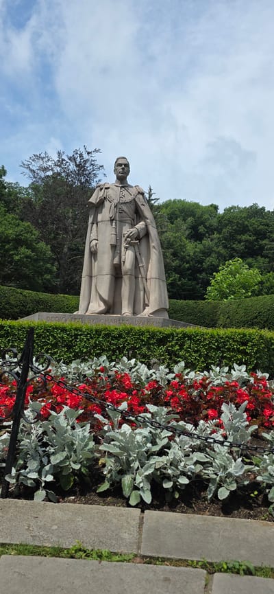 A stone statue of a man in regal attire stands in front of a dense hedge and lush green trees under a partly cloudy sky. The statue depicts a man wearing a long cape over a military uniform, complete with medals and a sword at his side. His hands are clasped in front of him. The base of the statue is inscribed with the words "DEDICATED BY CHARLES DALEY". In the foreground, a bed of bright red flowers is interspersed with silvery-green foliage, separated from the statue by a low, manicured hedge and a black metal fence with a chain. The statue is located in Niagara Falls, Canada, as evidenced by the inscription on the base.