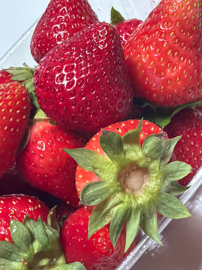 The image displays a close-up view of several fresh strawberries, likely in a clear plastic container. The strawberries are a vibrant red, with their characteristic tiny seeds visible on the surface. Some of the strawberries have green leafy tops, also known as calyxes. The lighting suggests an indoor setting, possibly a kitchen or a grocery store. There is no discernible location in Sauris / Zahre, Italy, or any indication of people or actions.
