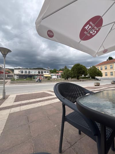 The image depicts a view from an outdoor cafe seating area in Garmen, Bulgaria, under a heavily clouded sky. In the foreground, a black rattan chair and a glass-topped table are visible. An open white umbrella with a red "Costa Coffee" logo is tilted overhead. Beyond the cafe area, a street runs horizontally across the frame, with a sidewalk and pavement in front of it. Across the street, several buildings are present, including a long, light-colored building with multiple windows, a yellow building with red trim, and other structures further in the distance. A few trees are scattered throughout the scene. Two individuals are seated on a low wall in front of one of the buildings. The overall atmosphere is overcast and slightly subdued.