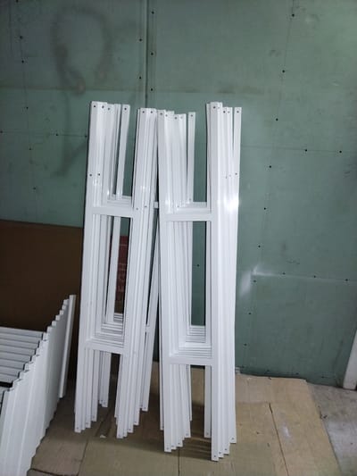 A group of white metal window frames are leaning against a green drywall wall in what appears to be a construction or storage area. The frames are rectangular with an opening in the center and appear to be of similar size and design. The wall behind them is painted green and has visible seams and circular indentations, possibly screw holes. The floor is covered with cardboard and concrete. To the left of the frames, a portion of a brown cardboard box is visible, with another white metal object with a ridged surface also leaning against it. The lighting is dim, casting shadows. There are no people or animals visible in the image. The overall impression is of an unfinished interior space.