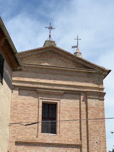 The image shows the facade of a brick building, likely a church, in Barbara, Italy. The building has a pitched roof with two crosses on top, one of which is topped with a flag. There is a single window on the facade, with a dark frame and multiple panes. The building is made of reddish-brown bricks laid in a stretcher bond pattern. A smaller, lighter colored building is partially visible on the left, with a gutter running along its edge. The sky is cloudy and blue. A bird is visible in flight in the lower right quadrant of the image. Electrical wires run horizontally across the facade.