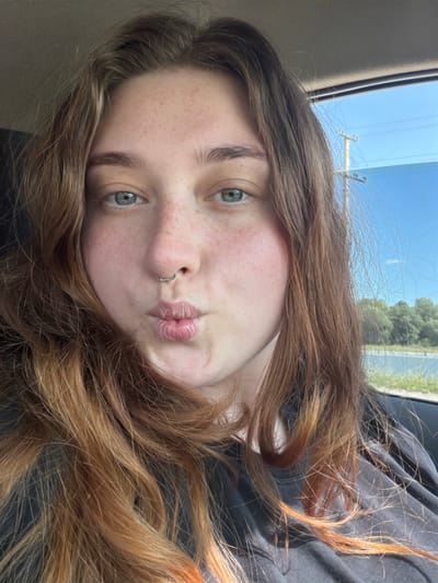 The image is a close-up selfie of a young woman with auburn hair, blue eyes, and freckles. She is wearing a dark t-shirt and has a silver septum piercing. She is puckering her lips and looking towards the camera. The background appears to be outdoors, with a blue sky and some trees visible through a car window. There is a utility pole and power lines in the distance. The image does not contain any information or context that would suggest the location is Duliby, Ukraine, or any other specific city.