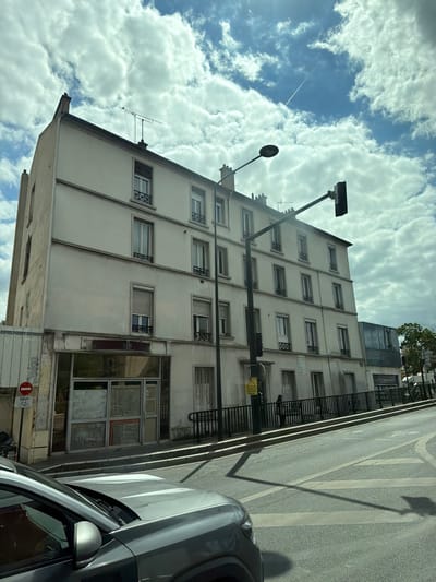 The image captures a street scene in Alfortville, France, focusing on a multi-story building and its surroundings. The building, with a pale facade, has several windows on each of its floors, some featuring decorative ironwork. The ground floor appears to be a commercial space with large glass windows, some of which are boarded up or obscured. A black metal railing runs along the front of the building at street level. A traffic light and a street lamp are prominently positioned in the foreground, casting shadows on the asphalt road. A gray car is partially visible in the lower left corner of the frame. The sky is filled with dramatic, white clouds against a blue sky, and a contrail from an airplane stretches across the upper right quadrant. A circular red and white sign with a bicycle symbol is affixed to a pole near the building's entrance. The overall impression is a somewhat overcast day in an urban environment.