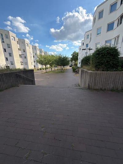 The image depicts an outdoor scene with modern apartment buildings on the left and a paved walkway on the right. The buildings are multi-story, with white facades and numerous square windows. Some of the windows have blue blinds visible. Green trees and bushes are interspersed between the buildings and along the walkway.
The walkway is made of dark gray paving stones arranged in a diagonal pattern. A low, concrete barrier runs alongside the walkway, with a bush growing over it. In the distance, a bicycle is leaning against the concrete barrier. The scene appears to be in a residential area.
The sky is bright blue with scattered white clouds, suggesting a sunny day. The time of day appears to be daytime, possibly late morning or afternoon, given the bright sunlight casting shadows. There is no visible text in the image. The overall mood is calm and ordinary, showing a typical day in a German urban or suburban environment. The perspective is slightly tilted, making the buildings appear to recede into the distance.