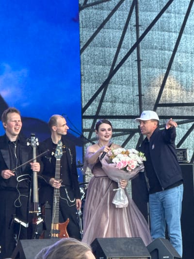 In Bolgar, Russia, on an outdoor stage with a blue backdrop and a metal scaffolding structure, a group of people is gathered. On the left, two men are positioned, one playing a cello and the other a bass guitar, both dressed in black suits. In the center, a woman in a light pink dress holds a microphone and a bouquet of pink and white flowers. To her right, a man in a dark jacket and a white baseball cap gestures with his right hand, seemingly addressing the woman or the audience. The stage is equipped with speakers and lighting equipment. The overall scene suggests a musical performance or award ceremony.