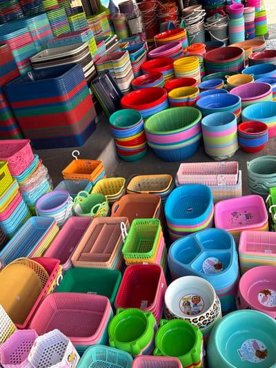 The image depicts a densely packed outdoor market stall in Ranong, Thailand, overflowing with a vibrant assortment of plastic household goods, predominantly containers of various shapes and sizes. The foreground is dominated by stacks of colorful plastic baskets and bowls, including large blue and red baskets, green colanders, and pink and blue pet bowls. In the background, rows of smaller, brightly colored containers are neatly stacked, creating a visually striking mosaic of plastic wares. The scene is brightly lit, suggesting it is daytime. There are no people visible in the image, but the arrangement of goods implies a retail setting. The ground appears to be paved, characteristic of a market street.