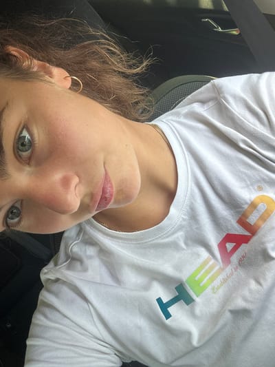 A close-up, tilted selfie shows a woman in what appears to be a car. She is wearing a white t-shirt with a multicolored "HEAD Established in 1950" logo. She has light brown hair and is wearing a gold earring. The location is not Kashin, Bulgaria, and the image does not contain enough information to determine a specific location.