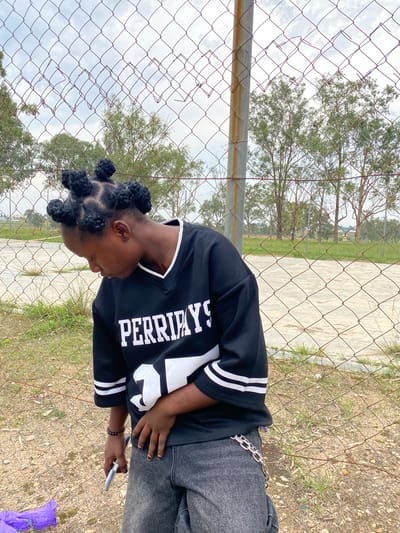 A young person with dark skin and braided hair styled into multiple spherical buns stands in front of a chain-link fence. They are wearing a black, oversized jersey with white lettering that reads "PERRYDAY" and white stripes on the sleeves. The jersey also has a large white number "31" on the front. They are also wearing dark, baggy jeans adorned with a silver chain belt. The person is looking down and holding a white object in their right hand, possibly a phone or cigarette pack. Their left hand is resting on their stomach. The background shows a cloudy sky, trees, a chain-link fence, and a paved area that appears to be a court or sports field. The ground is covered in dirt and sparse grass. The image is taken outdoors, likely in Zawan, Nigeria, given the context of the location.
