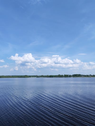 The image shows a body of water, likely a lake or river, with gentle ripples on the surface. The water appears dark blue, and the ripples create a textured pattern across the foreground. In the distance, a shoreline is visible, characterized by a line of green trees and some low-lying buildings or houses. Above the shoreline, a band of white, puffy clouds stretches across the sky, contrasting with the clear blue sky above. The scene is bathed in daylight, suggesting a sunny day. The city of Rechnaya Sosnovka, Russia is not discernible from the details in the image. There are no people or other actions visible in the image.