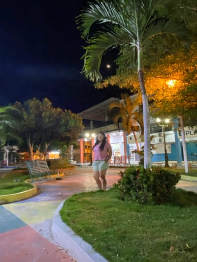 This image depicts a nighttime scene in a public park or square within General Villamil Playas, Ecuador. A single adult female stands in the center-right mid-ground, facing slightly left, with her hands in the pockets of her light green shorts. She wears a light pink short-sleeved top and appears barefoot. The environment features paved walkways with sections colored in pastel pink, yellow, blue, and green, bordered by manicured grassy areas and low shrubbery. Multiple park benches are visible, including a light green metal bench in the mid-ground. Several trees, including prominent palm trees, are illuminated by warm orange artificial light emanating from visible streetlights and building facades. In the background, multi-story buildings are discernible, one displaying a light blue facade with partial text "PLA" visible. The dark night sky in the upper right quadrant contains a distinct crescent moon.