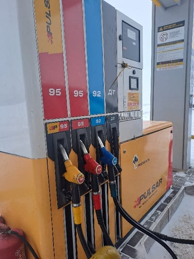 The image shows a gas station pump with four nozzles. From top to bottom, they are blue (92), red (95), and two yellow (95 and BP 95). Each nozzle is connected to a black hose. The pump has a digital display and price labels next to each nozzle. The structure is primarily gray, white, and yellow. Part of a fire extinguisher is visible at the bottom right of the image.