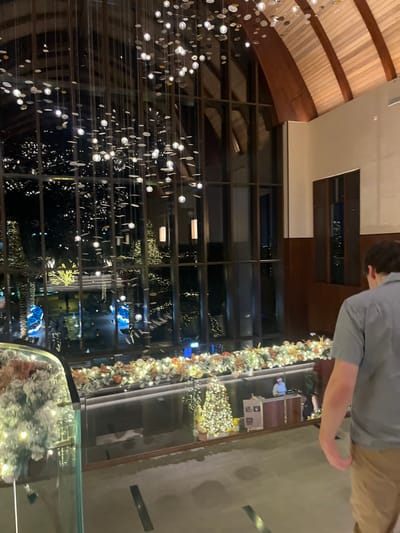 The image depicts the interior of the Ritz-Carlton Ras Al Khaimah, Al Wadi Desert, specifically its multi-level lobby area, viewed at night. The scene is dominated by a vaulted wooden ceiling with exposed beams and a large, multi-tiered chandelier composed of numerous illuminated disk-like elements. A vast glass facade on the left overlooks an outdoor area illuminated by countless small lights on trees and structures, suggesting a festive resort landscape with some blue-lit accents. The right wall features dark wood paneling and lighter cream sections.
In the foreground, partially visible on the left, is a glass railing adorned with a frosted, illuminated garland. A male subject with dark hair, wearing a light blue short-sleeved shirt and tan trousers, is seen from behind, walking towards the lower right of the frame. Below the observer's level, extensive holiday decorations are visible, including an illuminated garland spanning a lower railing and two decorated Christmas trees surrounded by wrapped presents. At a reception desk on the lower level, a person in a light blue uniform is seated, with a Santa Claus figure positioned on the desk nearby.