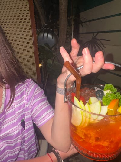 A medium close-up photograph taken at night in Ban Ko Li Pe, Thailand, shows an individual holding a large, clear glass beverage container. The person's right arm and torso are visible; they wear a light purple and white striped short-sleeved shirt with a dark fruit emblem on the left chest, a white beaded or ruffled neckline, a silver-colored mesh bracelet on the right wrist, and multiple dark beaded and red string bracelets on the lower right forearm.
The beverage container, resembling a fishbowl with a silver-colored wire handle, is filled with an orange-colored liquid and ice cubes. It is garnished with several slices of green apple, an orange segment, a sprig of green parsley, small dark berries (likely blueberries), and pieces of red fruit (likely strawberries). A brown plastic straw is inserted into the drink, held by the individual's right hand.
The background is dark, indicating a nighttime setting. Visible elements include dark tropical foliage, a light-colored woven rattan screen or partition on the left, and a dark, ribbed, inverted bowl-shaped object (likely a hanging light fixture or speaker) in the upper-center. A thin white cable runs vertically in the background. The scene depicts a leisure activity, likely at a restaurant or bar.