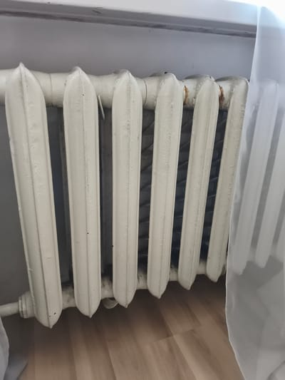 A close-up, eye-level shot shows a segment of an old, white cast-iron radiator situated against a light gray wall, with a wooden floor visible below. The radiator, composed of multiple vertical sections, exhibits signs of wear, including rust patches and chipping paint, particularly noticeable near the top. The lower portion of the radiator extends to the left, where a pipe fitting is visible. A sheer white curtain drapes over the right side of the radiator. The scene is consistent with an interior residential setting in Rahachow, Belarus, suggesting a typical dwelling with aging heating infrastructure.
