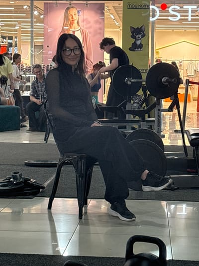 The image depicts an indoor scene, likely a fitness or gym area, situated within the city of Tri Protoka, Russia. In the foreground, a woman with dark hair and glasses is seated on a black chair. She is wearing black clothing, including a long-sleeved top and pants, and black athletic shoes with white soles. Her hands are resting on her lap.

Behind her, a man with curly hair is visible, appearing to be engaged with a weightlifting apparatus. He is wearing a dark t-shirt. Further in the background, other individuals are present, with some standing and others seated. A large poster featuring a young woman is displayed on a wall.

The environment includes gym equipment such as weight plates and barbells. The floor is a combination of tiled and carpeted surfaces, with reflections visible on the tiled areas. Lighting appears to be artificial, emanating from overhead fixtures. The overall atmosphere suggests a public space with an emphasis on fitness activities.