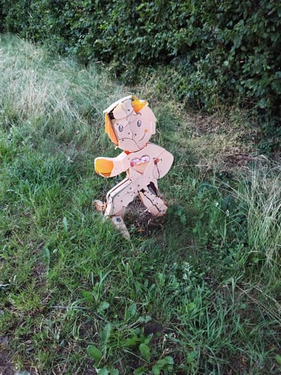 A pale, humanoid figure, possibly a scarecrow or a discarded toy, stands amidst tall grass and foliage. The figure is light tan with orange accents on its head and hands. It appears to be constructed from plastic or composite materials, with visible seams and cracks suggesting damage or wear. A heart shape is visible on its chest. The background consists of dense green bushes and dry grass. The scene is set outdoors, with the lighting suggesting it may be late afternoon or early evening. The location is identified as Schiffdorf, Germany.