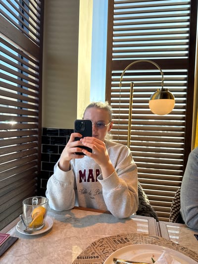 The image captures an indoor scene in the city of Mysiadło, Poland, set within what appears to be a restaurant or cafe. A young individual with light, curly hair and rimless glasses with a slight blue tint is seated at a light-colored, possibly marble, table. This person is wearing a grey crew-neck sweatshirt featuring a maroon graphic that reads "MAP Ville de Paris 1995". They are holding a dark-colored smartphone, consistent with an iPhone 14 Pro/Pro Max model, horizontally with both hands, positioned in front of their face as if taking a photograph or video of something to their right.
On the table to the left of the central figure, a clear glass mug with a handle contains a spiraled lemon peel and rests on a white saucer with a small spoon. Further to the right, a white plate with traces of green food residue and cutlery (a fork and knife) is present, situated on a round paper placemat printed with various Italian words such as "DELIZIOSO" and "PIZZA". A brass-colored lamp with a curved neck and a spherical white glass shade stands behind the table, to the right of the primary subject.
Natural light enters the space through a window, which is flanked by vertical wooden louvered blinds on both the left and right sides of the frame. A section of black tiled wall with white grout is visible behind the central figure's left shoulder. A second individual is partially visible on the far right, indicated by a grey-sleeved arm and a patterned chair.