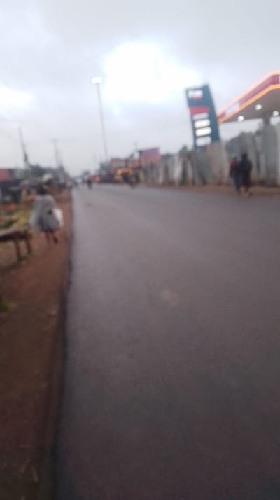 A blurry street scene shows a wet, gray asphalt road stretching into the distance under an overcast sky. A figure walks along the left side of the road. A gas station with a red roof and a tall sign are visible on the right, along with a few people. Other structures and vehicles appear indistinctly along the street. The image quality obscures details.