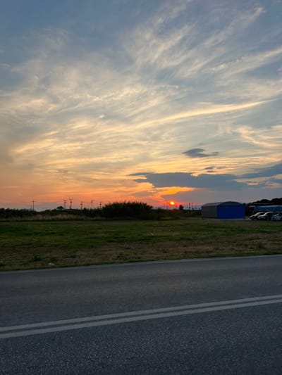 This is a photograph of a rural roadside scene at sunset in Chaidefto, Greece. The image is oriented vertically. The left side of the frame is dominated by a dramatic sky filled with wispy, elongated clouds that are illuminated by the setting sun. The colors in the sky range from a pale blue to warm oranges and yellows, indicating the golden hour.

On the right side of the frame, a paved road with two white lines runs parallel to a grassy verge. In the distance, beyond the grassy verge, a line of utility poles with wires is visible, suggesting the presence of infrastructure. Further in the background, silhouetted trees and what appear to be some distant hills or mountains can be seen. There are also a few structures visible, including a blue-topped portable building and some vehicles parked in the distance. The overall atmosphere is serene and peaceful, characteristic of a rural sunset. The time of day is clearly sunset due to the lighting and cloud colors. No people are visible in the frame. No text is visible.