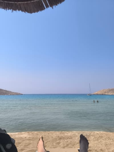 The image captures a serene beach scene on the island of Chalki, Greece, under a clear blue sky. In the foreground, two bare feet are visible, resting on the sandy shore, with one foot slightly closer to the viewer. A portion of a straw beach umbrella is visible at the top edge of the frame, casting a shadow. The beach transitions into calm, turquoise waters that extend to the horizon. Two rocky, sparsely vegetated hills flank the bay, framing the sea. A white sailboat with a blue sail is anchored in the distance, and several people are scattered throughout the water, some appearing to swim. The overall atmosphere is one of tranquility and relaxation, typical of a Mediterranean summer day.