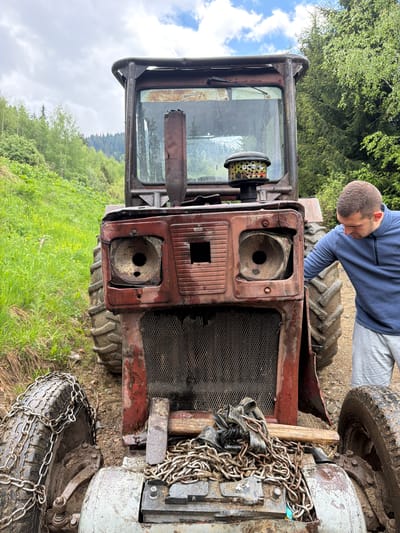 The image depicts a man leaning over the front of a weathered, red tractor. The tractor's headlights are missing, and the grill is visible. Chains are draped over the front of the tractor, near the wheels. The tractor is parked in a rural area with green vegetation and trees in the background. The setting appears to be in a mountainous region, possibly near a forest. Given the location hint, the scene is potentially located in the city of Рилски манастир, Bulgaria. The man is wearing a blue shirt and grey pants, and appears to be inspecting or working on the tractor.