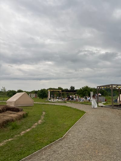 A wide shot captures an outdoor gathering on a cloudy day. In the foreground, a gravel path leads the eye towards a light-colored tent on the left, situated on a grassy field scattered with bales of hay. The path curves around the tent and heads towards a raised wooden platform on the right, adorned with string lights and supporting a wooden pergola.

Beyond the tent, several people are seated on hay bales and at tables, facing a stage-like area where a few individuals stand. Further back, more people are seen sitting in a larger audience area. A lone figure in a long white dress walks away from the camera on the gravel path. The background is filled with trees and a vast, overcast sky. The location is identified as Tavrichanka, Russia.
