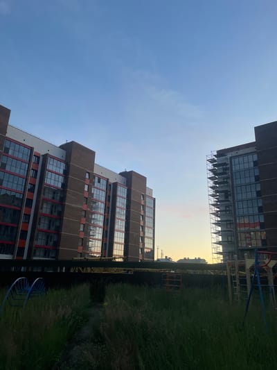 The image captures an outdoor scene with multiple tall buildings under a clear blue sky. The buildings are modern, featuring a repeating pattern of windows with dark frames and some red accents. One building, in the foreground and on the left, is viewed from a low angle, emphasizing its height. Another building, further in the background and on the upper right, appears to be under construction, with scaffolding visible on its exterior. Between the buildings, a playground structure with blue and yellow elements is partially visible, situated on a grassy area. The overall impression is that of an urban or suburban residential area, possibly a newly developed one, given the construction. The lighting suggests it might be late afternoon or early evening, with the sun casting a warm glow in the distance, particularly on the upper right building and the sky. The grass in the foreground is tall and somewhat unkempt. There are no people visible in the frame. No text is discernible. The location context provided is Laishevo, Russia.