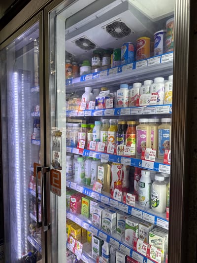 The image shows a refrigerated display case filled with various beverages, primarily milk and juice products, within a store located in Liwan District, China. The display case has glass doors with metal handles. On the right side of the doors, there's an orange sign with the Chinese characters "请拉门" (Please pull the door). The shelves are stocked with a variety of bottled and carton drinks, many of which have labels with Chinese text and prices displayed. Some shelves also feature fans for cooling. The lighting is bright, likely from within the display case and the surrounding store environment, highlighting the products on display. The overall impression is a well-stocked convenience store or supermarket chilled section.
