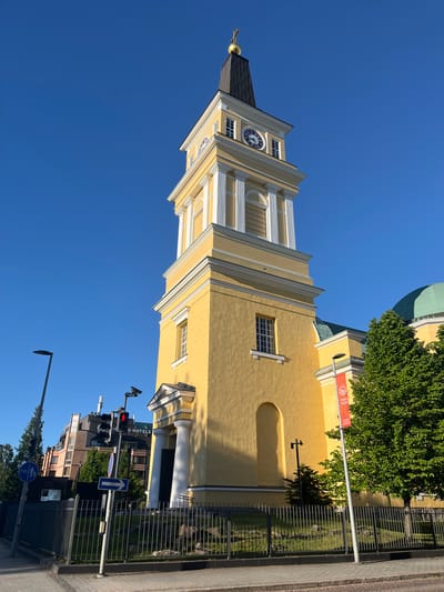The image shows a sunny day in Oulu, Finland, with a view of the Oulu Cathedral and its clock tower. The cathedral is painted a bright yellow with white trim and has a green dome visible to the right of the tower. The clock tower has a dark spire topped with a golden cross. A large clock face with Roman numerals is visible on the tower. In front of the cathedral, there is a metal fence and some greenery. To the left of the cathedral, there are modern buildings, street lights, traffic signals with a red light illuminated, and street signs. One sign indicates a bicycle path and pedestrian route, and another is a blue arrow pointing right. A red banner with "OULU 2026" is hanging from a pole in front of the cathedral. The sky is clear and blue.