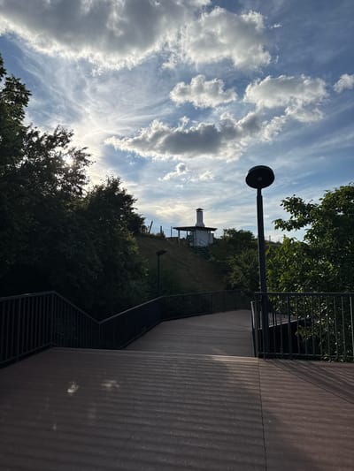This outdoor image, captured in Taman, Russia, depicts a contemporary pathway and its natural surroundings under a vibrant, partly cloudy sky. The scene is unpopulated, conveying a sense of serenity.

On the right side of the frame, a wide, dark-paneled structure, likely a wall or an elevated walkway, extends towards the background. Adjacent to it, a modern black metal railing runs alongside a long set of outdoor stairs, their dark treads leading upwards. A prominent black lamppost with a circular light fixture stands tall in the midground, its sleek form rising against the sky.

The midground and background feature a verdant hillside, richly covered with green trees and foliage. Sunlight, peeking through the dense canopy on the lower left, creates a bright lens flare, indicating a daytime setting, likely late morning or early afternoon. On the distant crest of the hill, a distinctive white structure with a dark, chimney-like element is visible, possibly a small building or a ventilation unit, with fences also apparent on the ridge. The upper portion of the image showcases a vast expanse of blue sky, dotted with bright white cumulus clouds and delicate, wispy cirrus formations. There is no discernible text within the image.