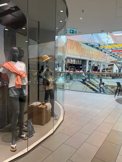 The image depicts the interior of a modern shopping mall, likely in Aachen, Germany, given the presence of a KFC outlet visible in the background. The scene is captured through a glass display window, showcasing mannequins dressed in casual attire.
In the foreground, two mannequins are displayed. The left mannequin is wearing a white t-shirt, denim shorts, and has an orange sweater tied around its shoulders. The right mannequin is wearing a striped top and brown shorts, with a straw hat. Below the mannequins, a wooden display box holds folded clothes. A price list is also visible in front of the mannequins.
In the background, several people are using escalators. Various retail outlets, including a KFC restaurant with signage indicating "Fresh all day", are visible. The mall features a contemporary architectural style with a high ceiling, modern lighting fixtures, and decorative elements on the walls. The floor is tiled. The overall atmosphere suggests a typical bustling shopping environment.